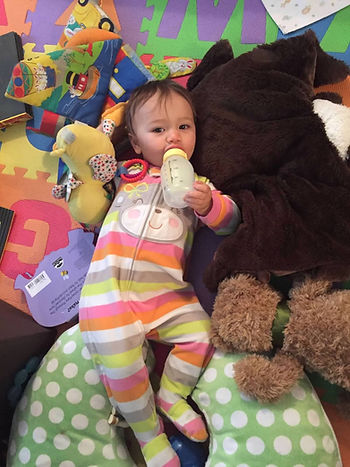 Baby in striped onesie lies on colorful play mat, holding a bottle. Surrounded by plush toys and cushions, creating a cozy scene.