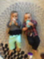 Two babies lie on a patterned rug, drinking from bottles. One wears a striped shirt and teal pants, the other a plaid shirt and jeans.