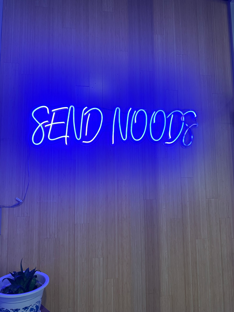 Blue neon sign on wooden wall that says 'Send Noods'