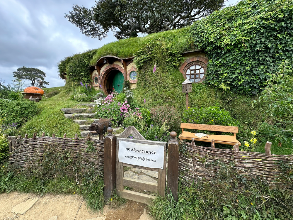Bilbo Baggins's Hobbit Hole in Hobbiton Grass-covered Hobbit house with a round door, colorful flowers, wooden bench. Sign reads "no admittance except on party business." Serene mood.