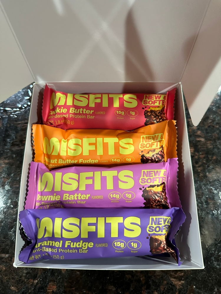 Misfits Protein Bars Box of MISFITS plant-based protein bars in colorful wrappers displayed on a dark marble surface. Flavors: Cookie Butter, Peanut Butter Fudge, Brownie Batter, and Caramel Fudge.