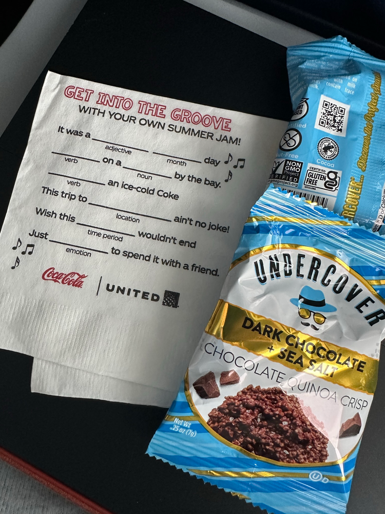 Undercover Dark Chocolate + Sea Salt Chocolate Quinoa Crisp Napkin with a summer-themed fill-in-the-blank activity, featuring Coca-Cola and United logos. Nearby is a blue pack of chocolate quinoa crisps.