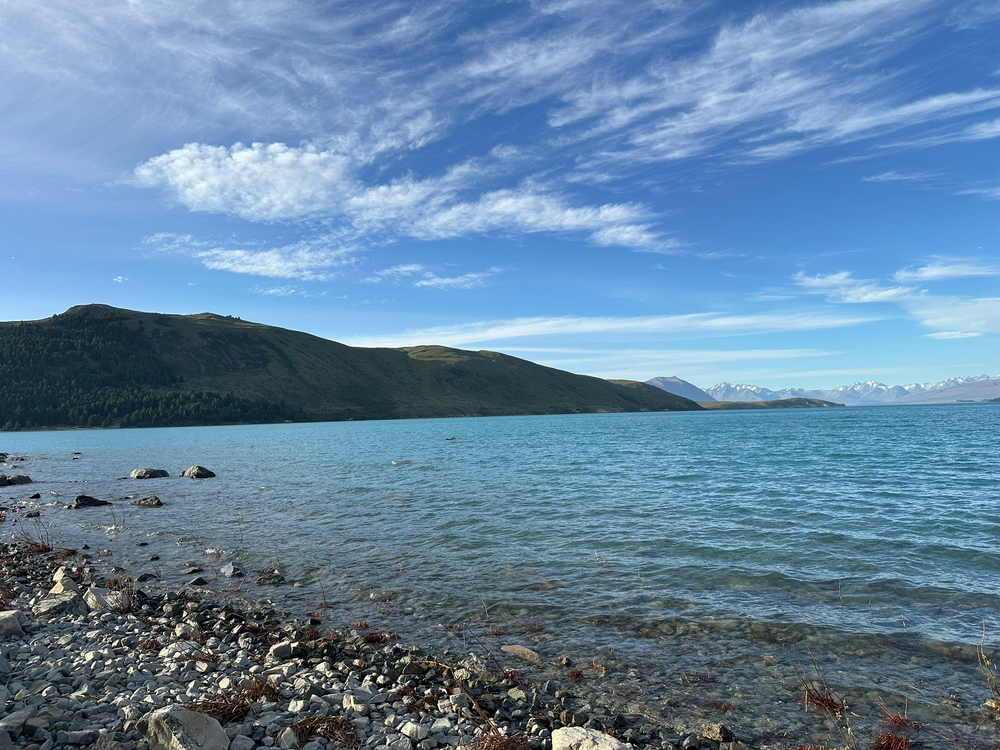 Calm lake with rocky shore, clear blue water, and distant green hills. Fluffy white clouds scattered across a bright blue sky.