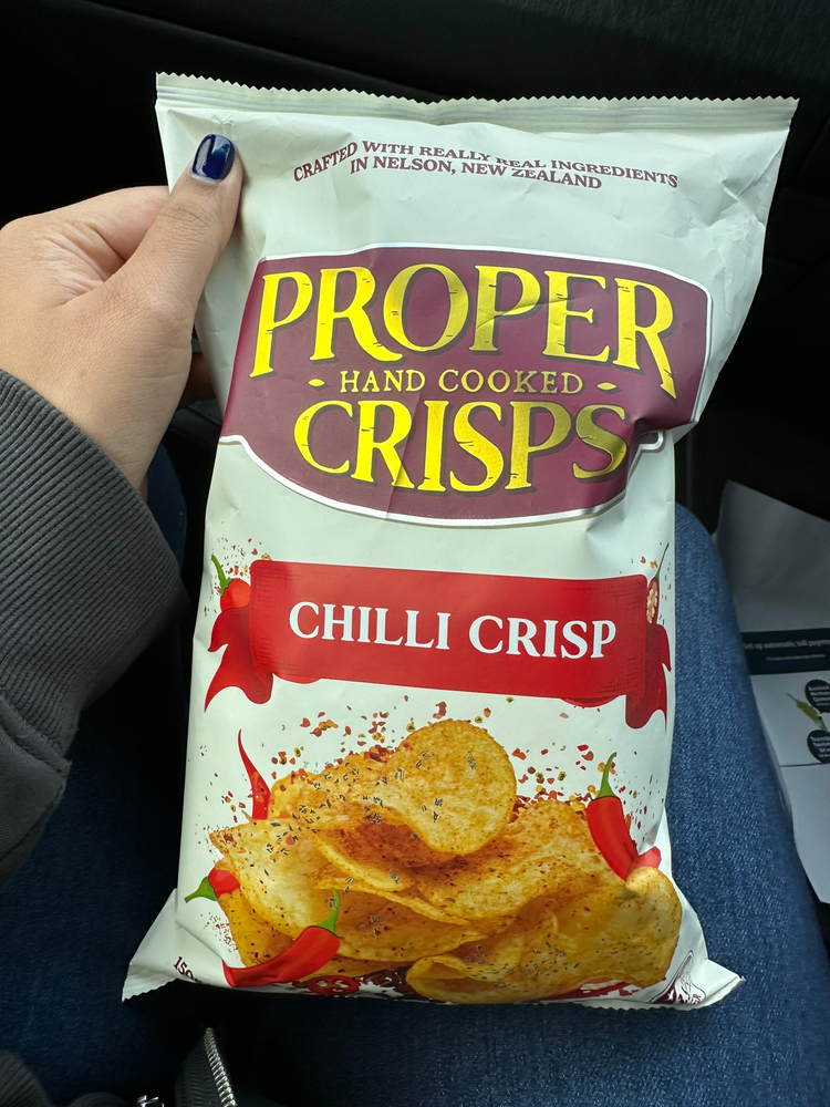 Chilli Crisp-flavored Proper Crisps Hand holding a bag of Proper Crisps Chilli Crisp flavor. The setting appears casual, with the blue fabric of jeans in the background.