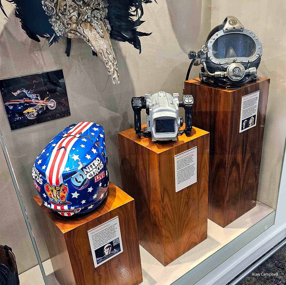 Display case with three items. A red, white and blue motocross helmet, underwater camera and diving helmet.