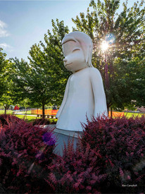 Very large white sculpture of what looks like an Asian child, sunlight shining through trees behind.