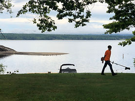 Man walking on grass with trimmer by lake; overcast sky.