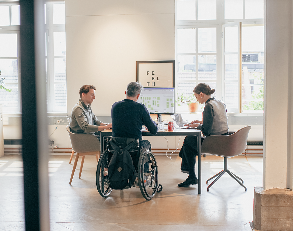 Three people in an office meeting at a table, one person uses a wheelchair, bright natural light from large windows, landscape photo.