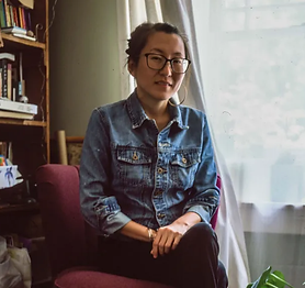 Image shows organizer and writer Mia Mingus sitting comfortably in her living room. She wears a denim jacket, glasses, and hoop earrings.