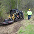 Two workers clearing brush on a hillside. One is operating a skid steer with a brush mower and the other is walking beside it and is wearing a neon green shirt and yellow hard hat