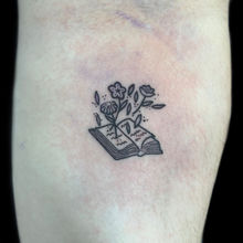 Minimalistyczny tatuaż z motywem książki, z której wyrastają kwiaty. Mały, graficzny wzór w stylu ignorant, wykonany w studiu Hypnotic Tattoo w Bielsku-Białej.