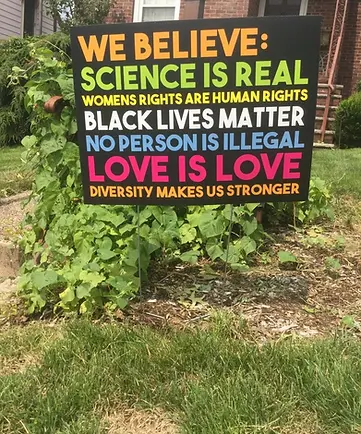 Colorful lawn sign that reads, We Believe: science is real, women's rights are human rights, Black lives matter, no person is illegal, love is love, diversity makes us stronger. Placed on a green and brown lawn.