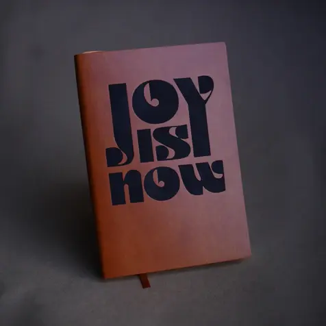 A photograph of a brown faux leather journal with the words "Joy is Now" on the cover