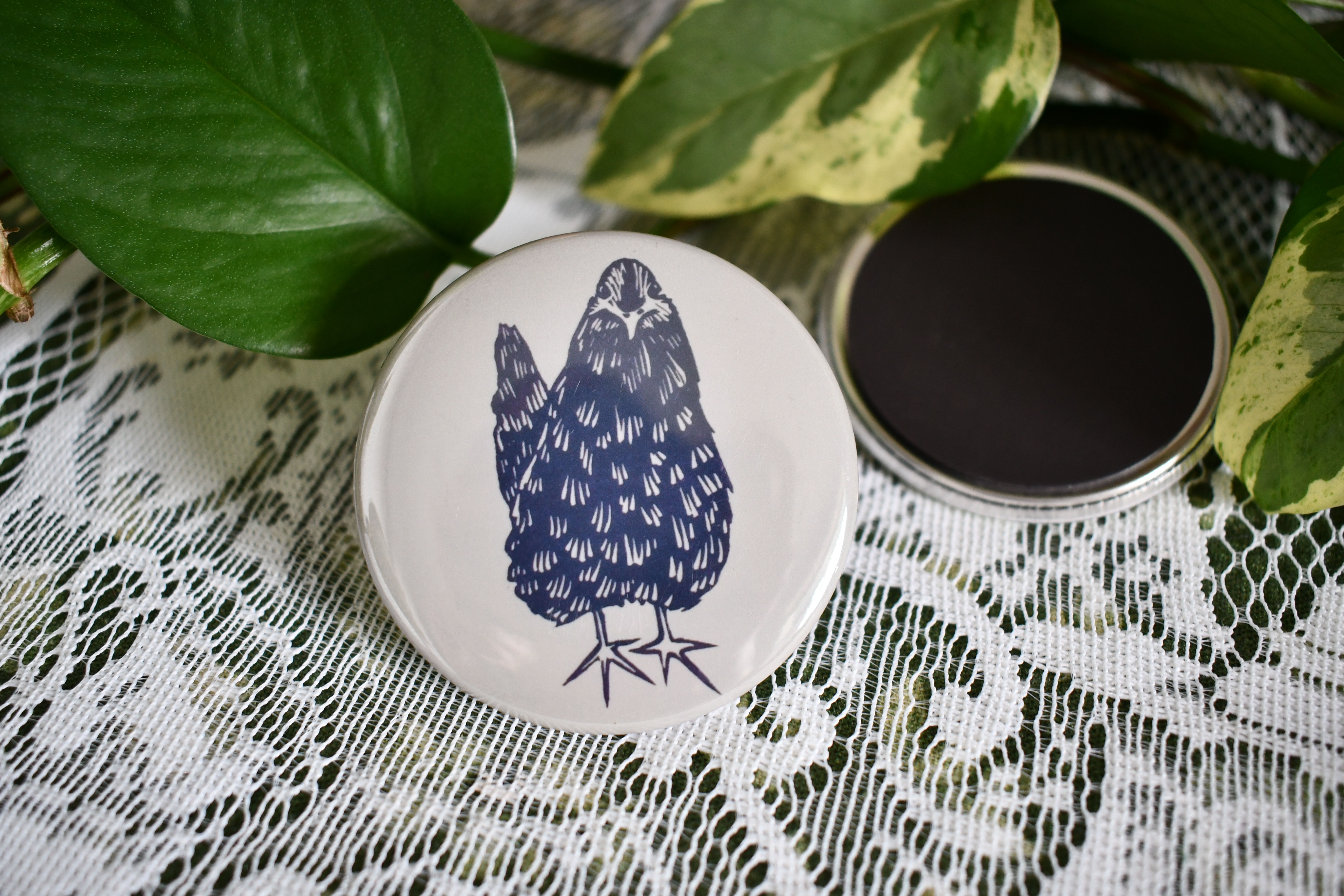 White hen button with a blue drawing, sitting with green leaves backdrop.