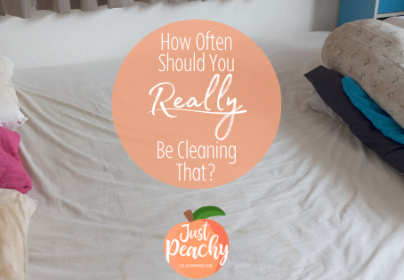Messy bed with multiple pillows and blankets. Text overlay reads: "How Often Should You Really Be Cleaning That?" with a "Just Peachy Cleaning Co." logo.