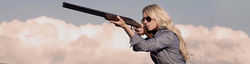 Pretty blonde girl with shotgun & shooting glasses, taking aim with blue sky & cloud background.