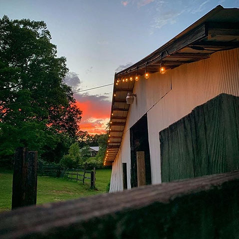 The barn at the center of Pastor Zac's family's farm. A white barn with cafe lights strung along the eves. The sun sets behind clouds and trees in the background.