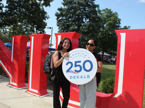 Two students hold the America 250 DeKalb logo in front of large NIU letters.