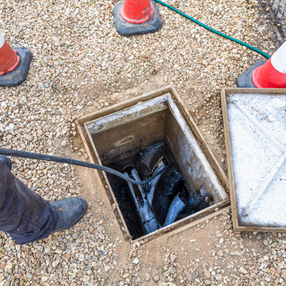 Worker inspecting a manhole with tools, open lid, gravel surroundings, Clear Choice Pressure Washing Portugal