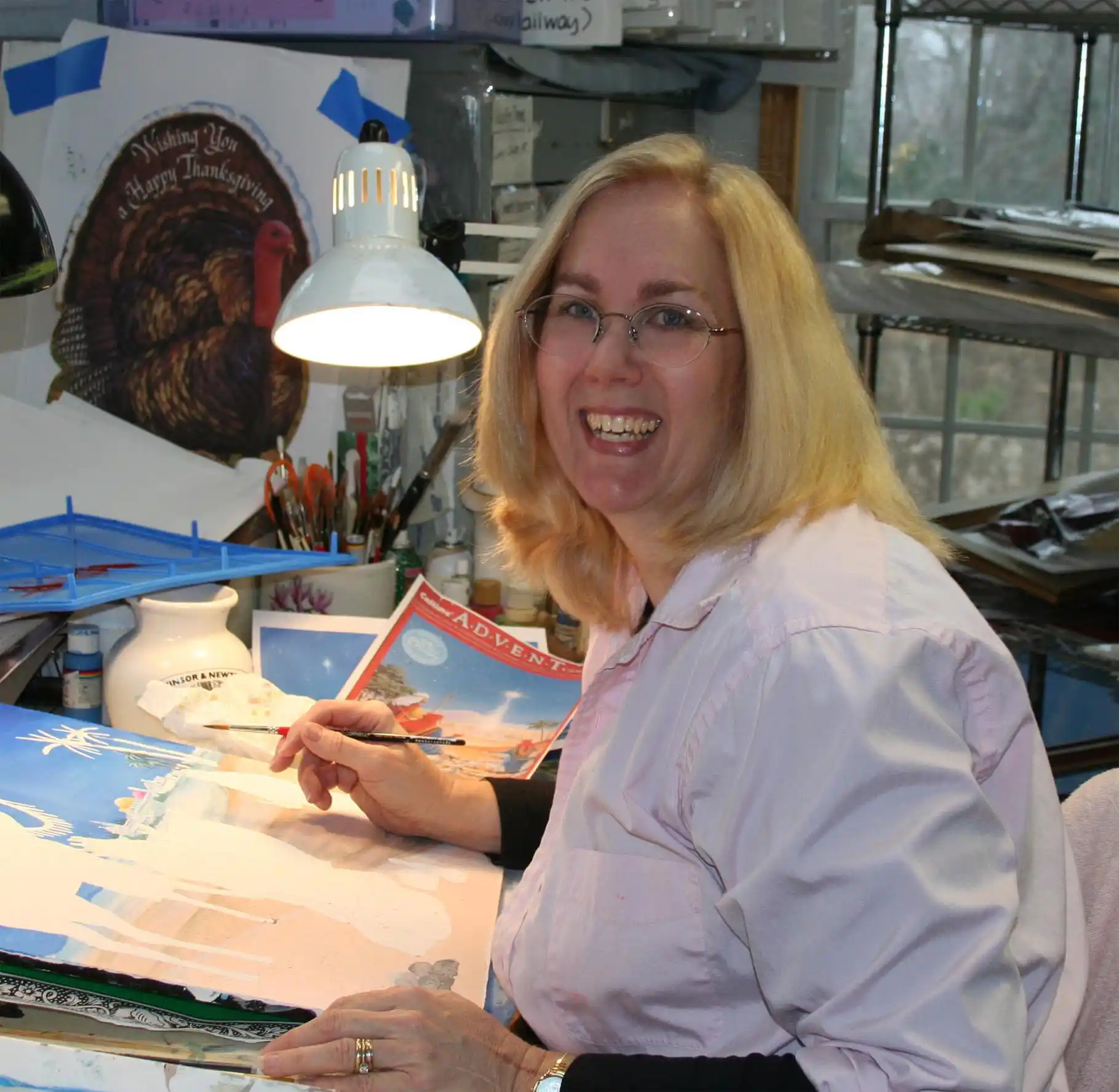 An image of the artist, Mary Lou Troutman. A woman with blonde hair and glasses sits in an artist studio, working on a painting of Southern Maryland. A variety of supplies surround her, including brushes, mats, painters tape, and works in progress.