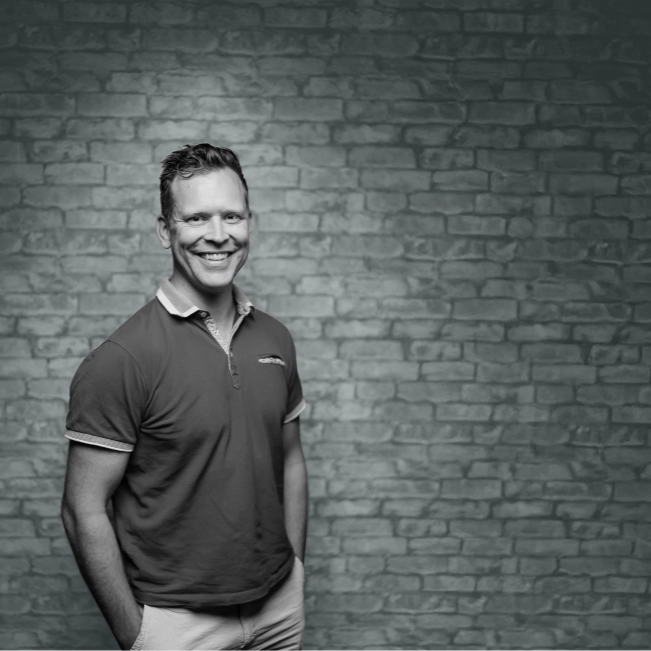 A man with short hair stands smiling in front of a brick wall.