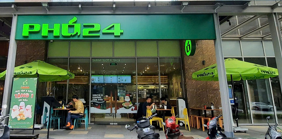 People dine outside a restaurant named "Phở 24" under green umbrellas. The setting is urban, with scooters parked nearby.