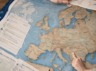 Hands pointing at a detailed map of Europe, with documents beside it. The setting is a wooden table, creating an analytical mood.
