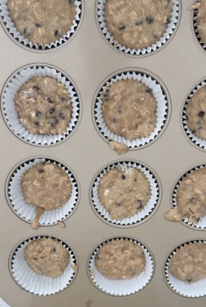 Tan muffin tin filled with high protein banana oat muffin batter, sprinkled with salt, cinnamon, walnuts, and brown sugar.