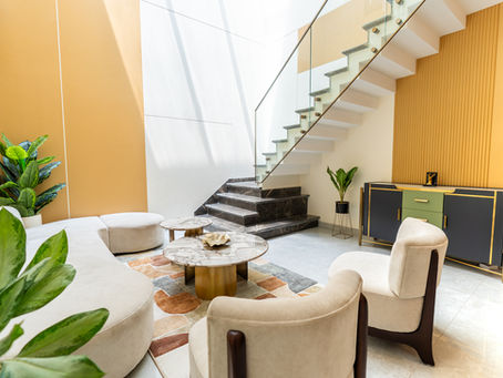 Modern living room with beige sofas, marble tables, staircase, and plants. Orange accent wall and colorful rug create a cozy, elegant vibe.