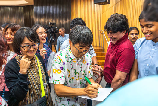 Man signing autographs surrounded by excited fans at an event with smiles.