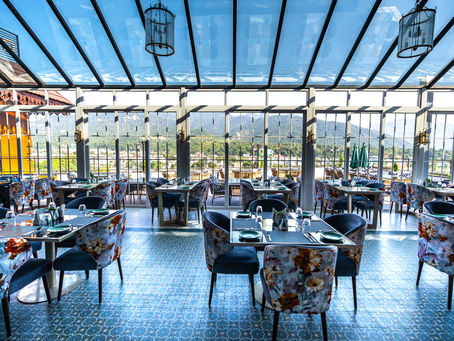 Elegant restaurant with floral chairs and blue tiles under a glass roof. Mountain view outside the windows, creating a serene ambiance.