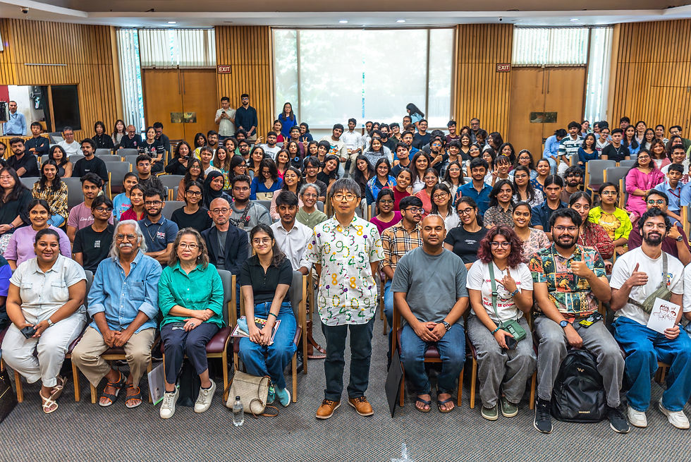 A large group of people seated in an auditorium, with one person standing at the center. Bright, casual attire; wood-paneled background.