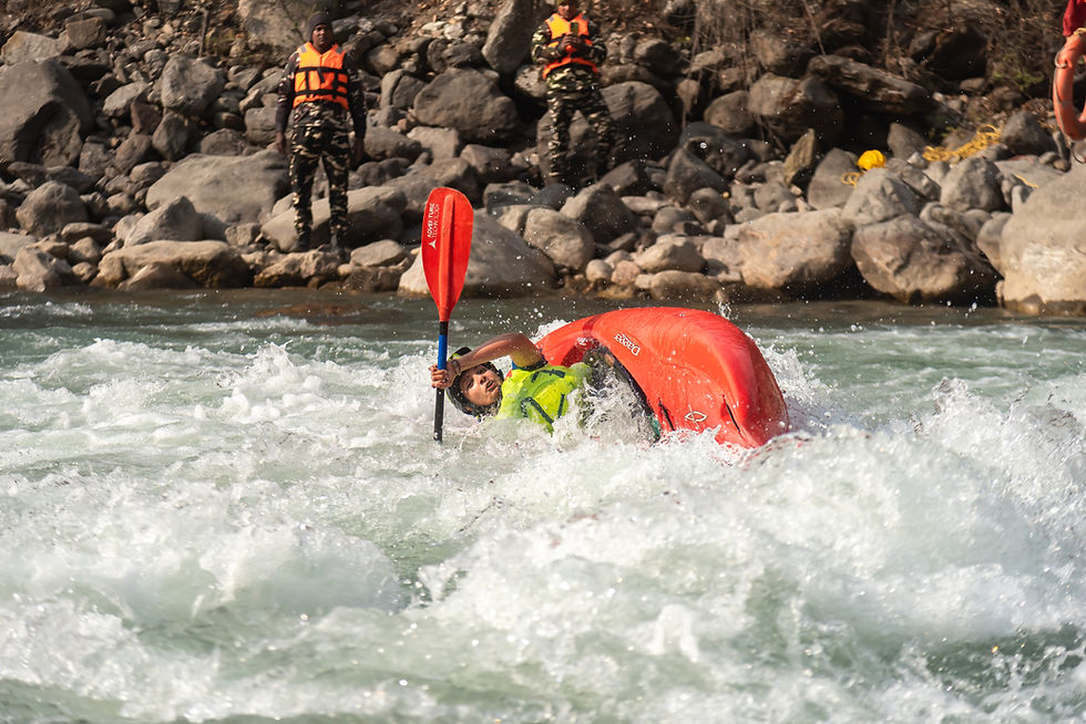 Kayaker in a red canoe navigates white-water rapids, paddling vigorously. Two observers in orange vests stand on rocky riverbank.
