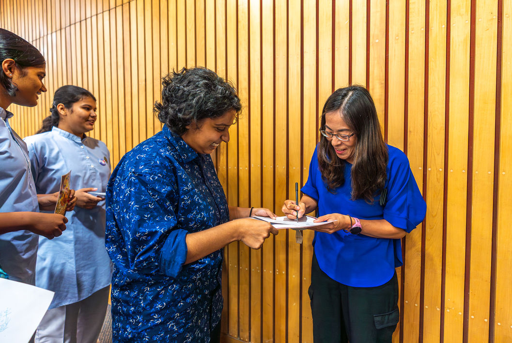 Two women signing documents, smiling in front of a wood panel background.