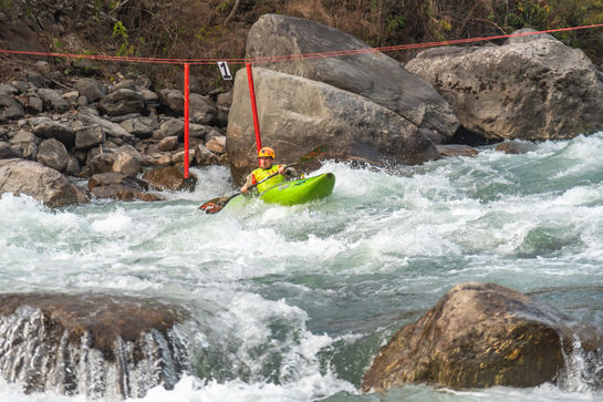 Kayaker navigates rapids, wearing helmet and life vest, with red markers in background.