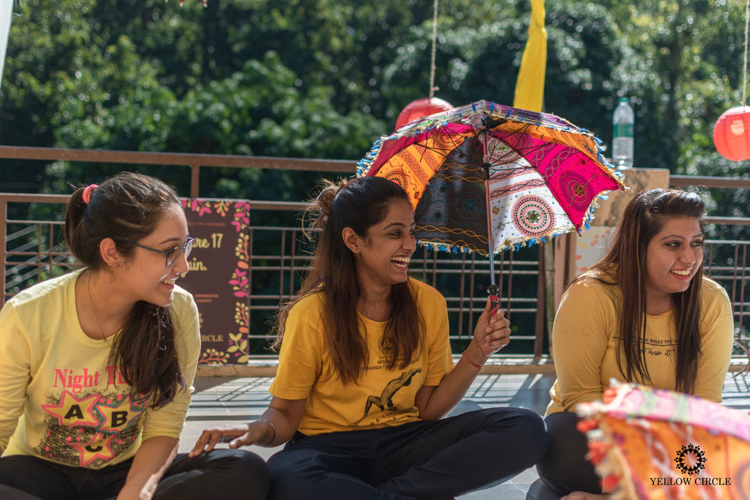 A social event photograph of three women laughing; one holds colorful umbrella outdoors with trees in the background.