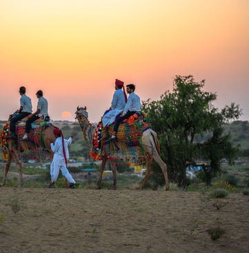 People riding camels at sunset in a desert landscape with colorful decorations.