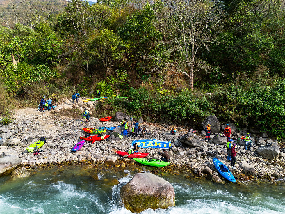 People in colorful gear prepare kayaks on a rocky riverbank. Lush trees in background. A banner reads "START" nearby. Energetic mood.