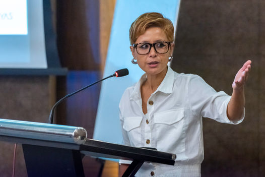 Woman speaking at a conference, wearing glasses and a white button up shirt.
