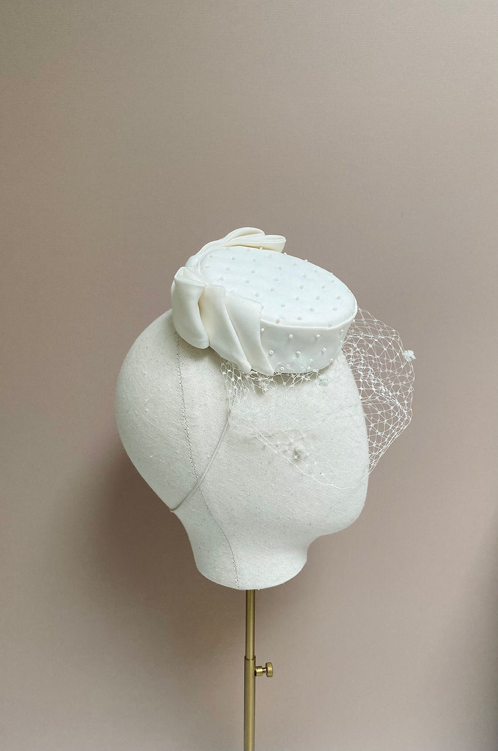 Thumbnail: An ivory pillbox hat with veiling, bow and bead decoration is displayed on a mannequin head