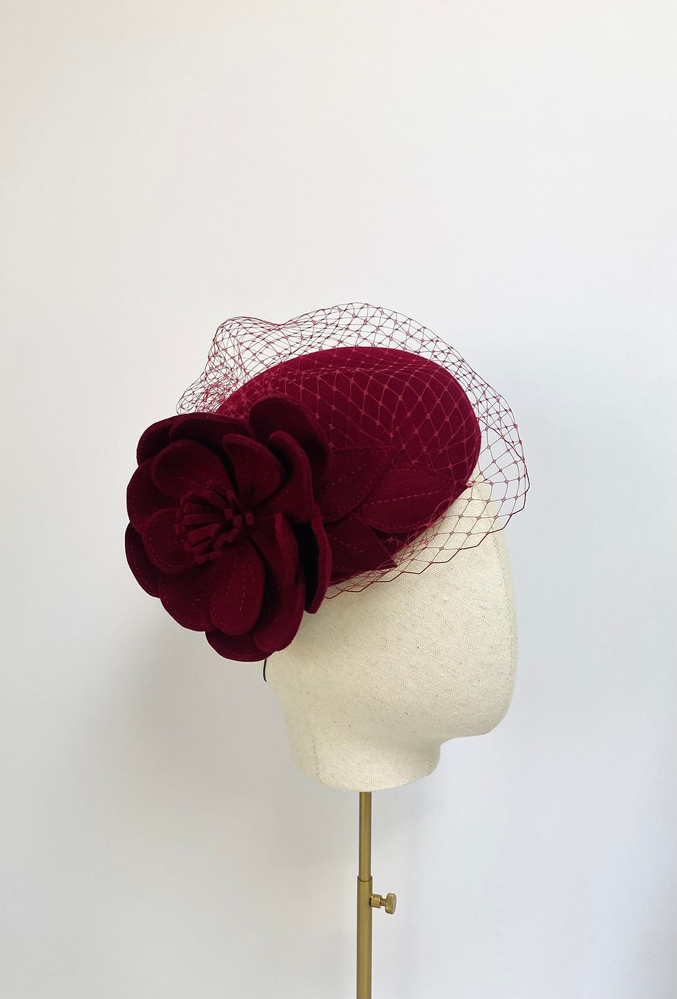 A dark red felt flower wedding hat with a veil is displayed on a mannequin head