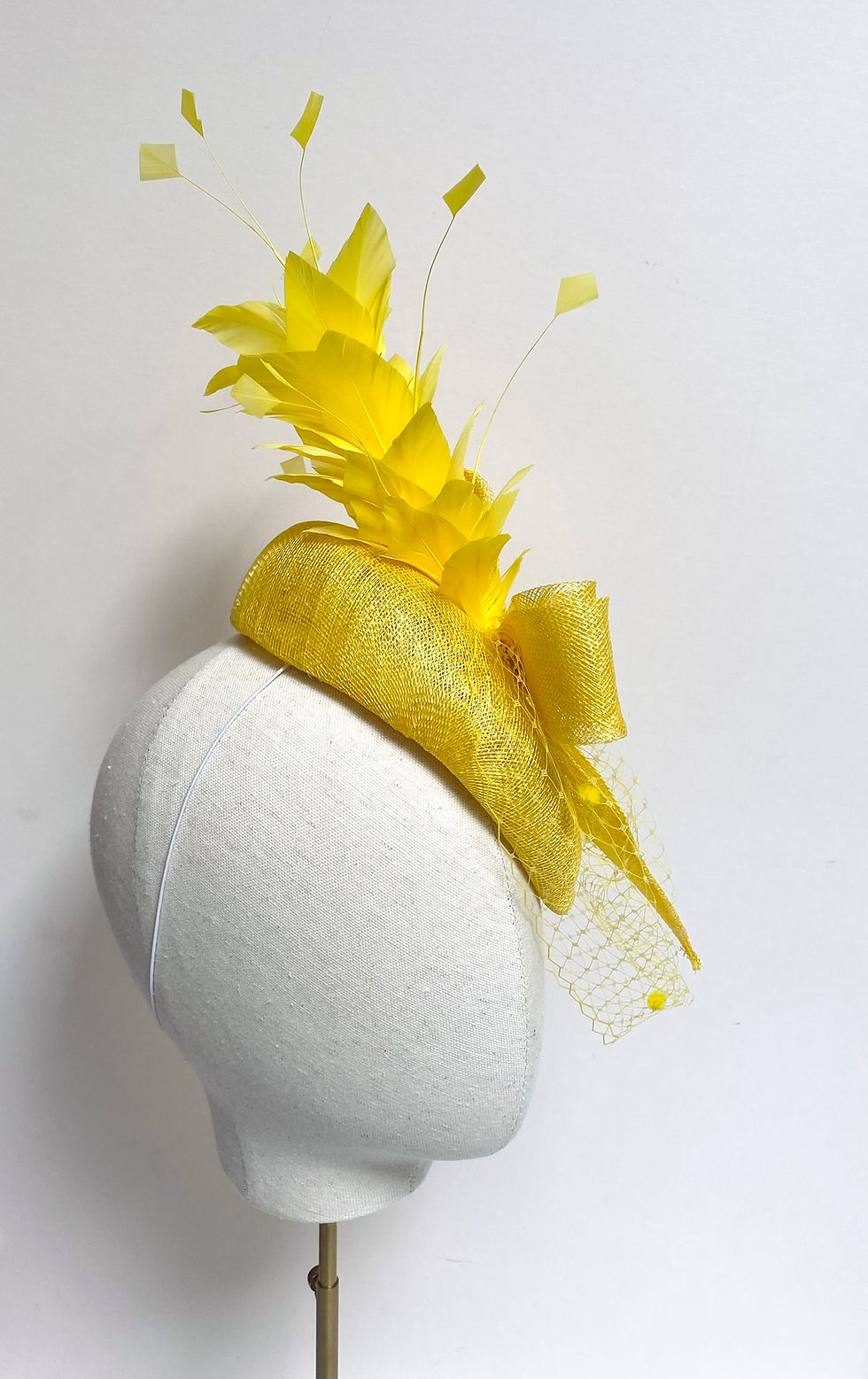 Thumbnail: A yellow feather fascinator with spot veiling and matching bow is displayed on a mannequin head
