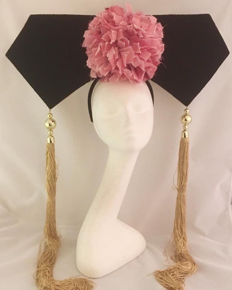 A large flat black headpiece with a pink pom pom and tassels is displayed on a mannequin head.
