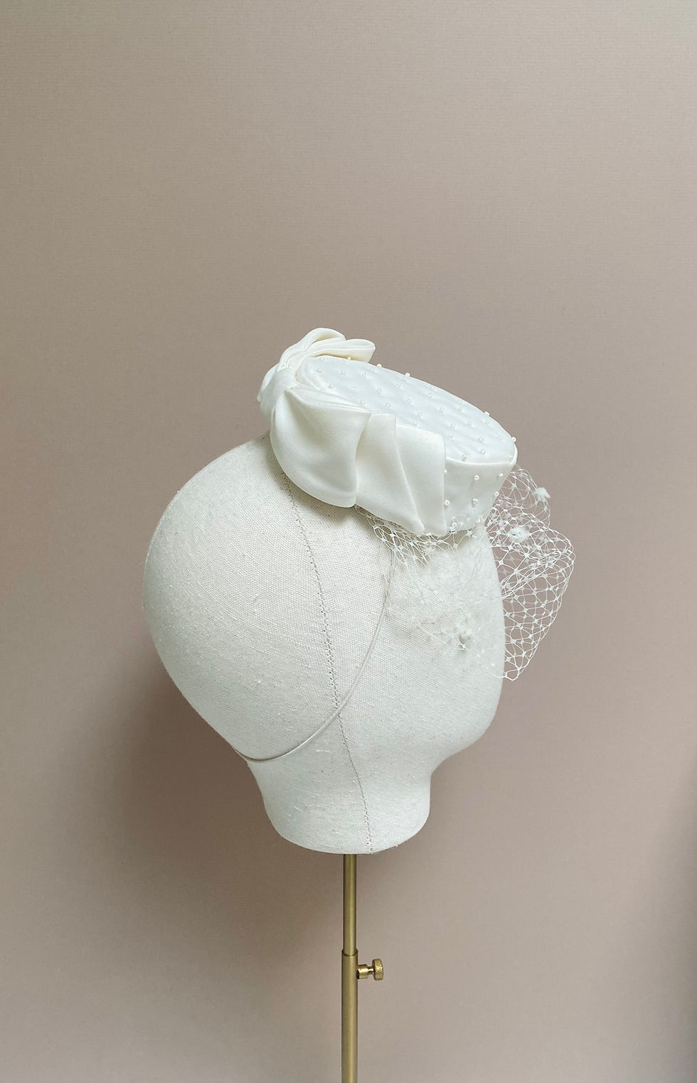 Thumbnail: An ivory pillbox hat with veiling, bow and bead decoration is displayed on a mannequin head