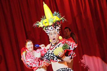 A man wearing a fruit headdress and colourful outfit is stood against a red curtain.