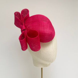 A bright pink teardrop shaped hat with a large double bow is displayed on a mannequin head