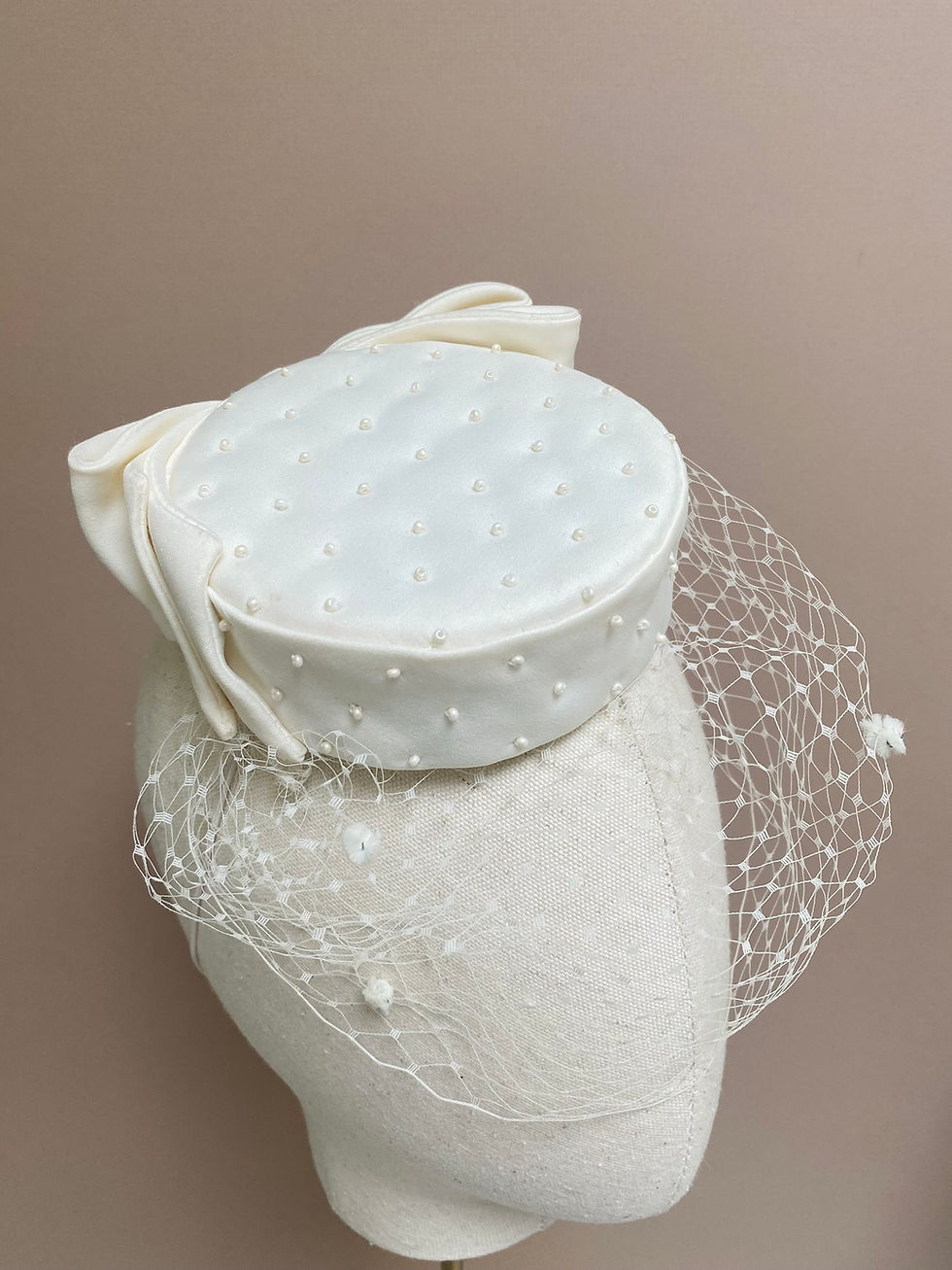 Thumbnail: An ivory pillbox hat with veiling, bow and bead decoration is displayed on a mannequin head