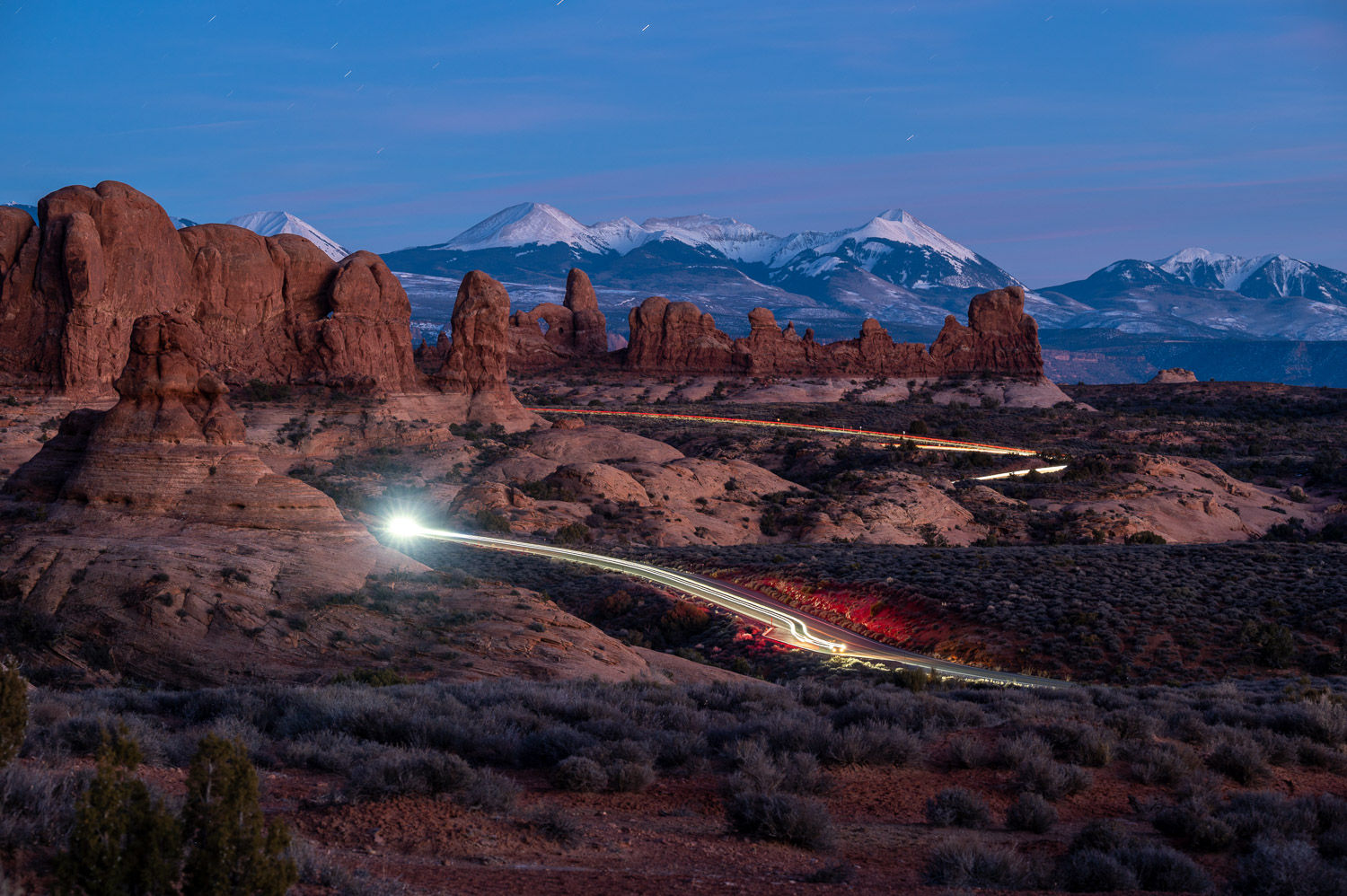 Blue hour landscape in Arches National Park, Utah with glowing sandstone formations, a winding road, and a car light trail un
