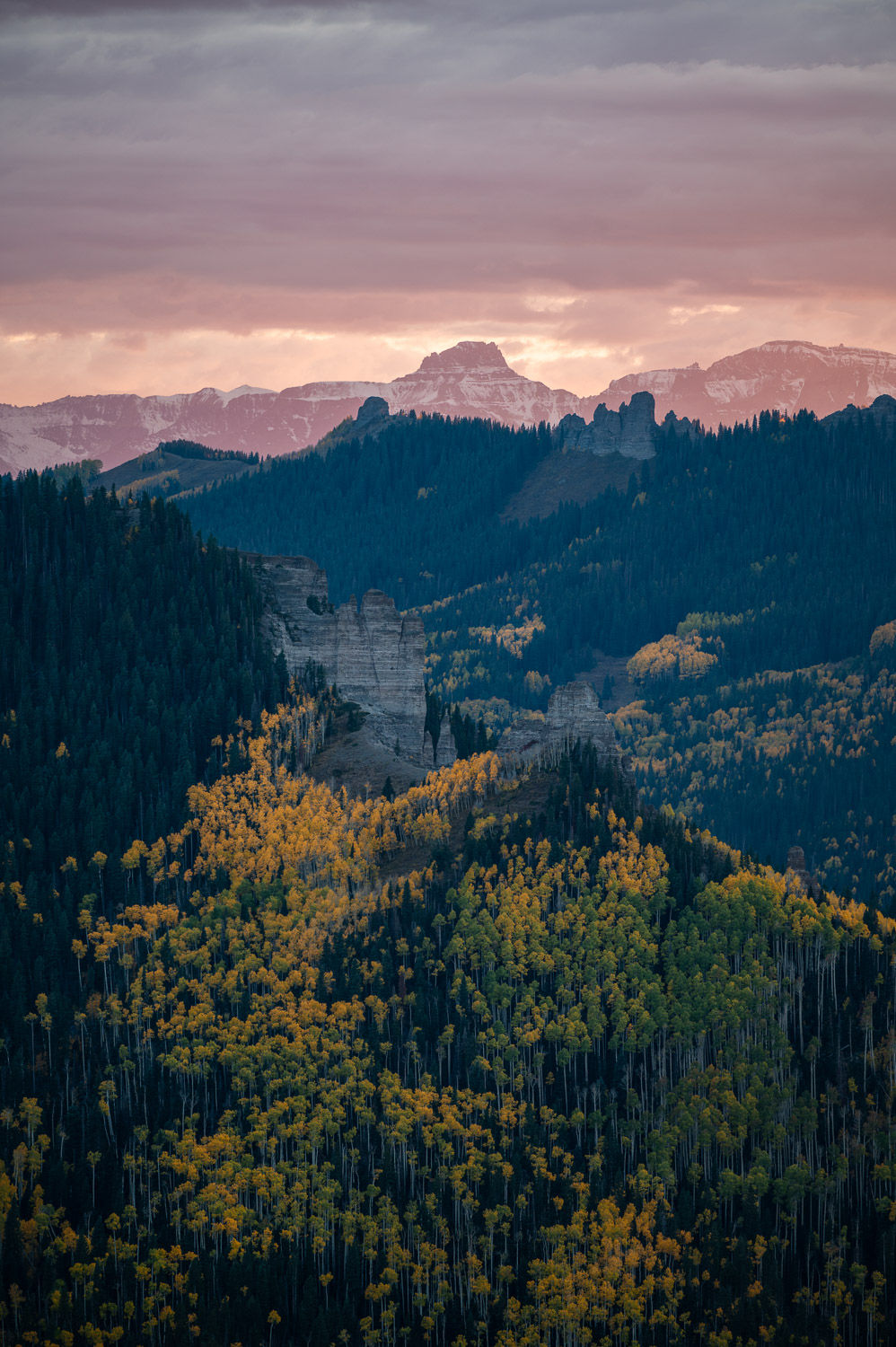 Golden aspen-covered hillside with patches of green, layered mountain ridges, and distant peaks under a warm pink sky in Sout
