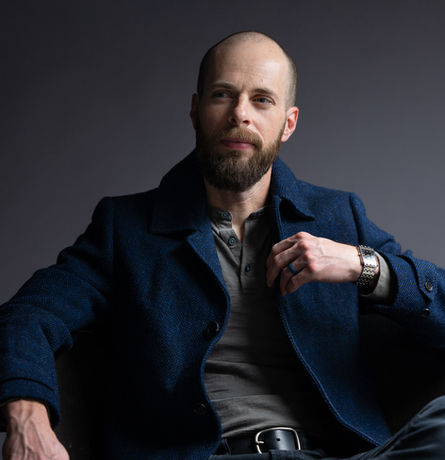 Dramatic studio portrait of a bearded man in a textured blue coat seated in a chair, styled for creative headshots and editorial profiles.